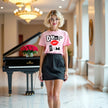Full body shot of female model wearing light pink “Dump Him” tee with bold letters and red lips standing at piano bar.